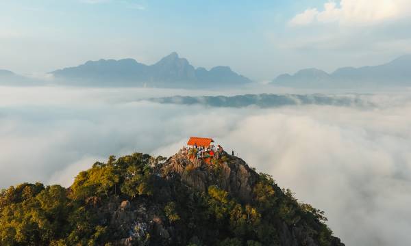 Eine grüne Bergkuppe ragt in die Wolken, auf ihrer Spitze ein rotes Wellblechdach, daneben eine Gruppe Menschen, die in die Ferne schaut.