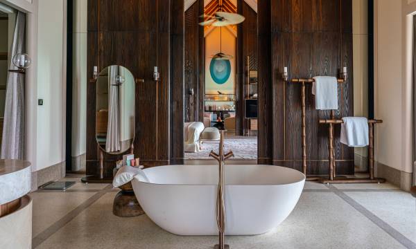 Eine freistehende Badewanne mit goldener Armatur in einem Raum mit hellem Marmorboden und edler Holzvertäfelung an der Wand. In einem Spiegel hinter der Badewanne spiegelt sich ein luxuriöser Raum mit einem Bett.