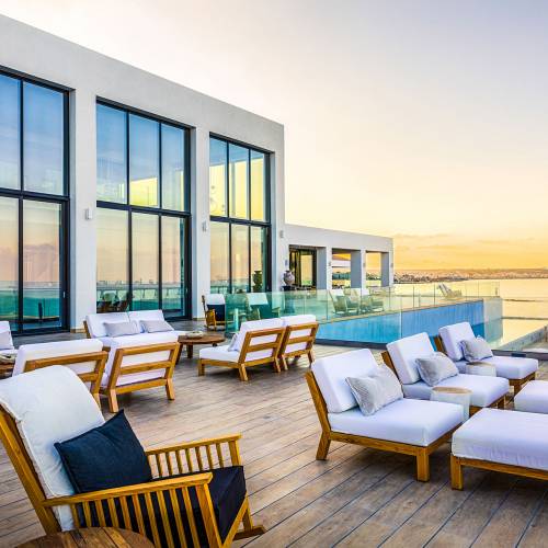 Terrasse einer modernen Hotelanlage mit Holzliegen und weißen Auflagen, Blick auf das Meer und den Sonnenuntergang, große Fensterfront.
