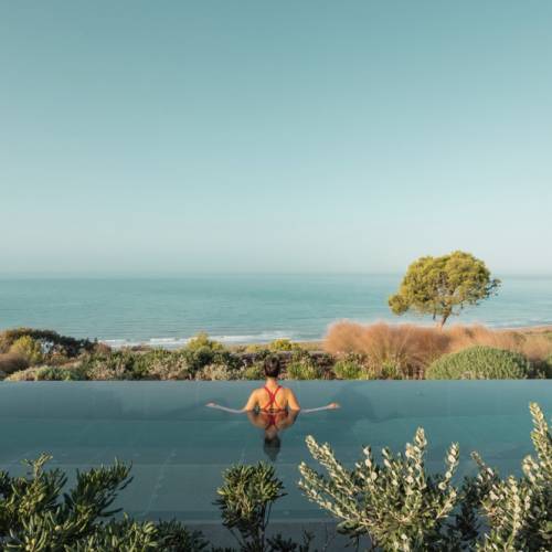 Person im roten Badeanzug entspannt im Infinity-Pool mit Blick auf das Meer und Küstenlandschaft