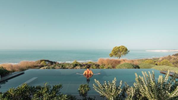Person im roten Badeanzug entspannt im Infinity-Pool mit Blick auf das Meer und Küstenlandschaft