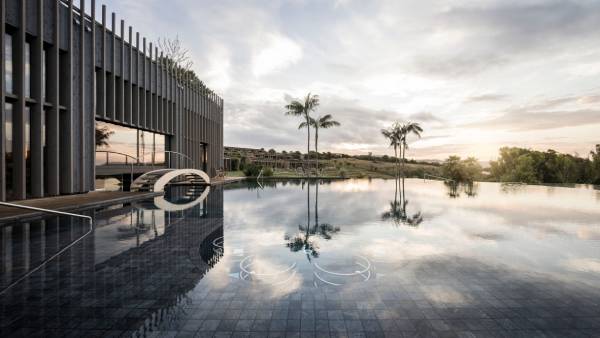 Moderner Infinity-Pool mit reflektierender Wasseroberfläche neben Gebäude mit vertikalen Lamellen und Palmen im Hintergrund