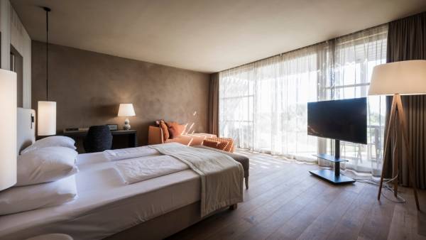 Hell gestaltetes Hotelzimmer mit Doppelbett, Schreibtisch, orangefarbenem Sessel und großem Fenster mit Vorhängen