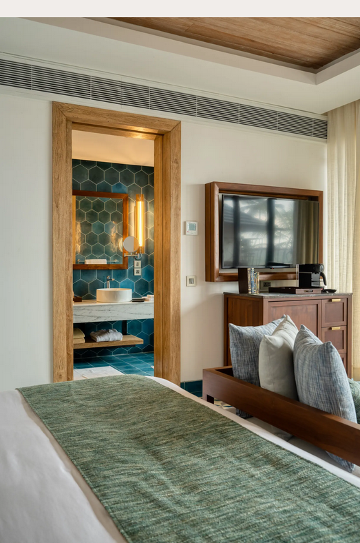 Hotelzimmer mit Bett und grüner Tagesdecke, Blick in ein Badezimmer mit blau-grünen Fliesen und Waschbecken, Fernseher auf Kommode