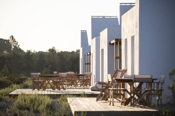 Moderne Hotelanlage mit weißen Gebäuden und Holzterrassen. Auf den Terrassen stehen mehrere Holzstühle und Tische. Im Hintergrund sind Bäume sichtbar.