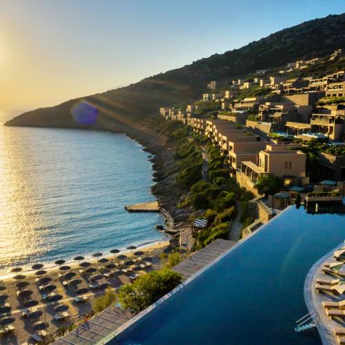 Luftaufnahme einer Hotelanlage am Meer mit mehreren Gebäuden auf einem Hang, einem Infinity-Pool und Liegen am Strand. Sonnenuntergang im Hintergrund.