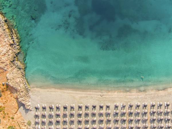 Luftaufnahme einer Hotelanlage am Strand mit mehreren Sonnenschirmen in Reihen auf dem Sand. Das Wasser ist klar und türkisfarben.