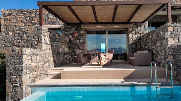 Hotelanlage mit einem Poolbereich, umgeben von Steinmauern. Zwei gepolsterte Stühle und ein Tisch stehen auf der Terrasse. Ein Treppeneingang führt zum Pool.