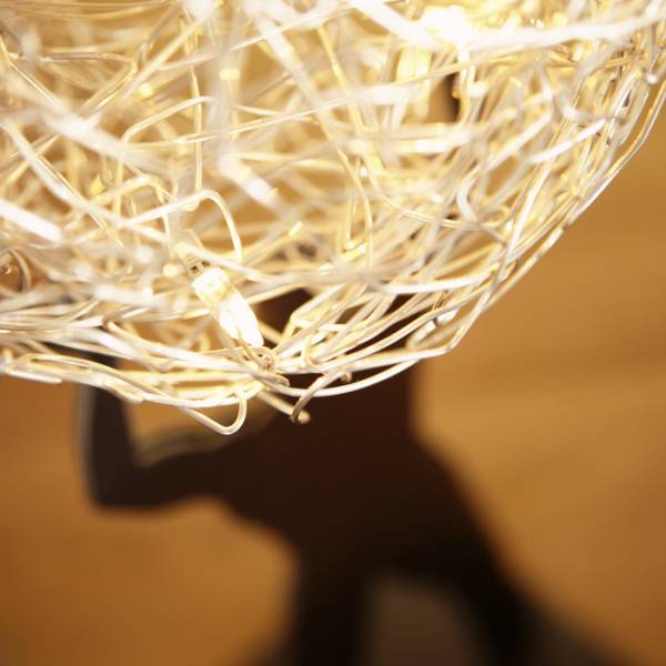 Detailaufnahme einer modernen, kunstvollen Lampe mit einem Drahtgeflecht, das warmes Licht abstrahlt. Im Hintergrund ist der Schatten einer Person zu sehen.