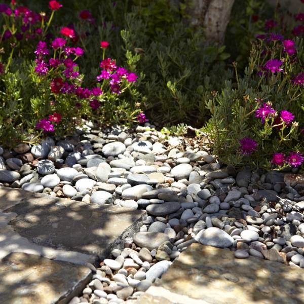Gartenansicht einer Hotelanlage mit bunten Blumen und Kieswegen. Sichtbare runde Steine und quadratische Platten im Vordergrund.