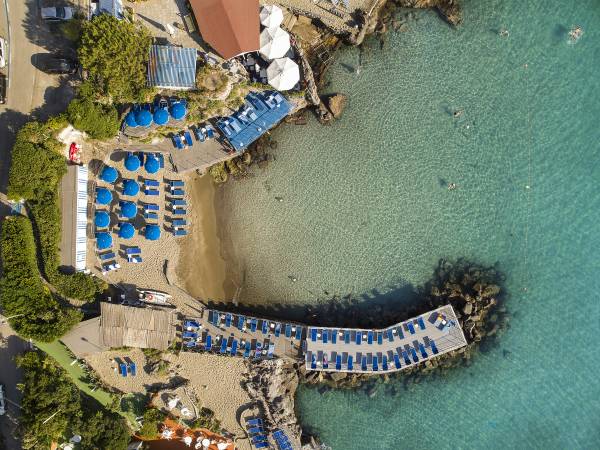 Luftaufnahme eines Strandes mit blauen Sonnenschirmen und Liegen, Steg mit weiteren Liegen, klares Meerwasser und einige Schwimmer