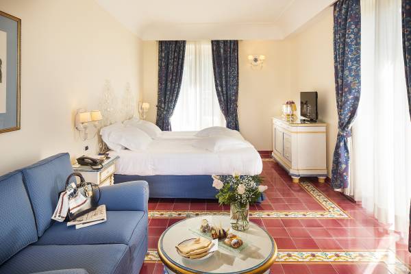 Hotelzimmer mit Doppelbett, blauem Sofa, rundem Glastisch mit Blumenstrauß und Snacks, helle Wände und große Fenster mit blauen Vorhängen
