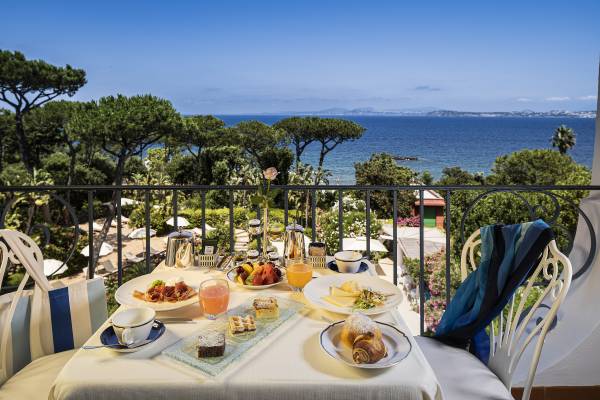 Frühstückstisch mit Croissant, Käse, Obst, Saft und Kaffee auf Balkon mit Blick auf Meer und Bäume