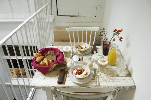 Ein gedeckter Tisch mit einem weißen Stuhl, darauf befinden sich eine Schüssel mit Joghurt, frisches Obst, Croissants in einem Korb, ein Glas Orangensaft und eine Kanne. Im Hintergrund ist eine Treppe und eine weiße Wand sichtbar.