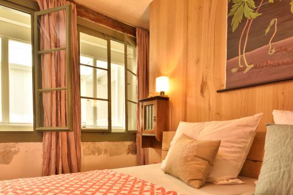 Ein Hotelzimmer mit einem Bett, weißen Kissen und einer dekorativen Decke in Korallenfarbe. An der Wand hängt ein Bild mit Palmen. Fenster mit grünen Rahmen und Vorhängen.