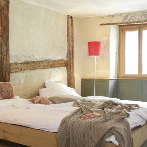 Ein helles Hotelzimmer mit einem großen Holzbett, weißen Bettlaken und einer grauen Decke. Eine rote Stehlampe steht neben dem Fenster. Im Hintergrund ist eine Person in Bewegung.