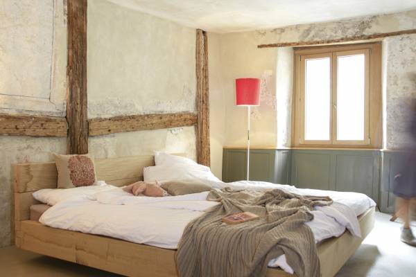 Ein helles Hotelzimmer mit einem großen Holzbett, weißen Bettlaken und einer grauen Decke. Eine rote Stehlampe steht neben dem Fenster. Im Hintergrund ist eine Person in Bewegung.