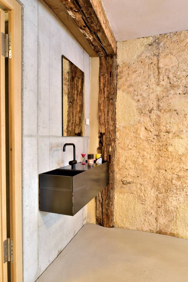 Moderne Badezimmeransicht mit einem schwarzen Waschbecken und einem Spiegel an einer Wand aus Beton und Holz. Auf dem Waschbecken stehen Pflegeprodukte.