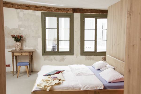 Helle Hotelzimmeransicht mit einem Holzbett, zwei Kissen, einem Tisch mit einer Vase und einem Stuhl. Fenster mit grünen Rahmen.
