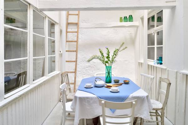 Ein Tisch mit blauer Tischdecke steht in einem hellen Raum. Auf dem Tisch befindet sich eine weiße Teekanne, eine kleine Schale und ein Glas mit Blumen. Um den Tisch stehen vier Stühle.