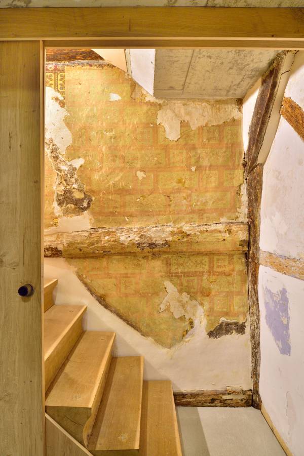Blick auf eine Treppe aus Holz, die zu einem Raum führt. An der Wand sind beschädigte Tapeten und freiliegende Balken sichtbar.