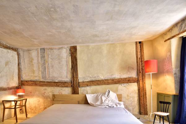 Hotelzimmer mit einem großen Bett, einem Nachttisch mit roter Lampe, einem Stuhl und einer Kommode. Sichtbare Holzbalken an der Wand.