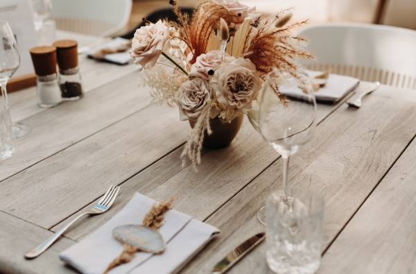 Tischgedeck in einem Restaurant mit einem Blumenarrangement aus rosa und beige Rosen sowie Trockenblumen in einer Vase. Besteck und Gläser sind ordentlich platziert.