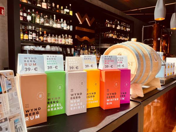 Sechs Flaschen der Marke 'Wyndberg' stehen auf einem Tisch. Die Flaschen sind in verschiedenen Farben und enthalten Rum, Apfelbrand, New Make Spirit, Winter Gin. Im Hintergrund sind Regale mit Spirituosen und ein Holzfass sichtbar.