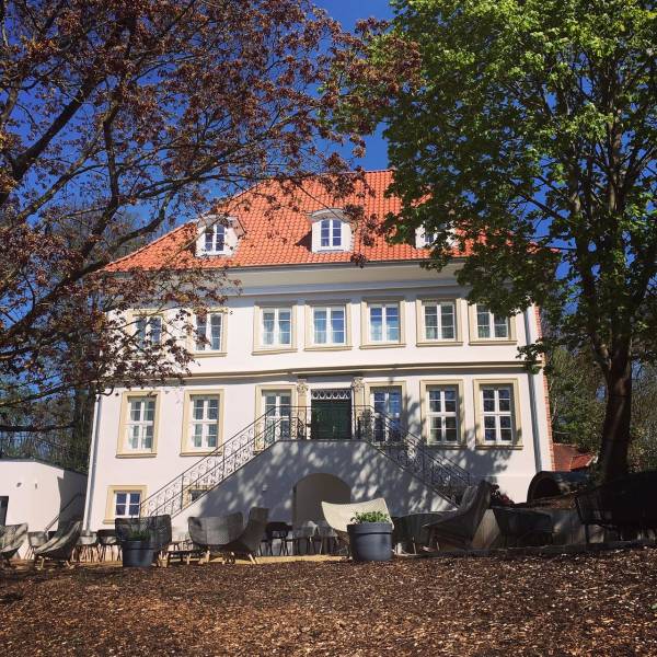 Hotelanlage mit einem dreigeschossigen Gebäude, rotem Satteldach und weißen Wänden. Vor dem Gebäude befinden sich Terrassenmöbel und große Pflanzenkübel.