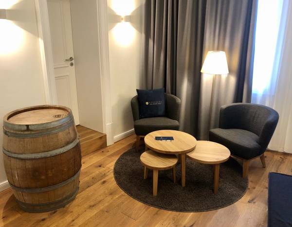 Hotelzimmer mit zwei grauen Sesseln, einem runden Holztisch und einem Holzfass als Tisch. Eine Stehlampe und ein Teppich sind sichtbar.