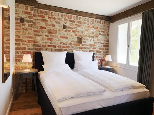 Doppelzimmer in einer Hotelanlage mit zwei Betten, weißen Bettdecken und Nachttischen. Sichtbare Ziegelwand und Fenster mit Vorhängen.