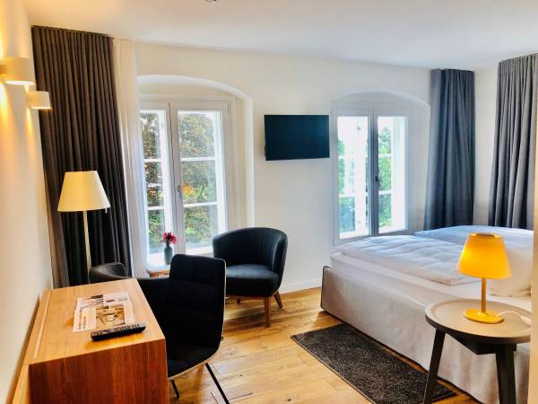 Hotelzimmer mit einem Doppelbett, einem schwarzen Sessel, einem Schreibtisch aus Holz und einem Fernseher an der Wand. Fenster mit Blick auf einen Garten.