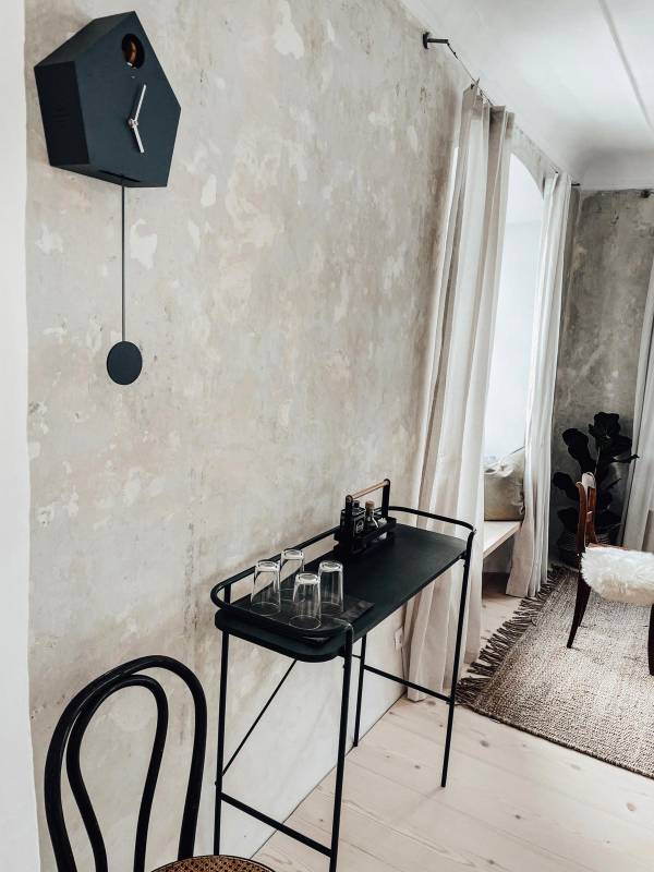 Ein minimalistischer Raum mit einer grauen Wand. An der Wand hängt eine graue Wanduhr in Form eines Vogelhauses. Auf einem schwarzen Tisch stehen vier Gläser und eine kleine schwarze Schreibmaschine. Ein Stuhl aus Holz ist sichtbar.