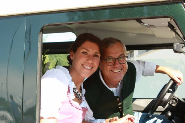 Ein Paar in traditioneller Tracht sitzt im Fahrerraum eines grünen Fahrzeugs. Die Frau trägt ein Dirndl mit einer Halskette, der Mann trägt eine Weste.