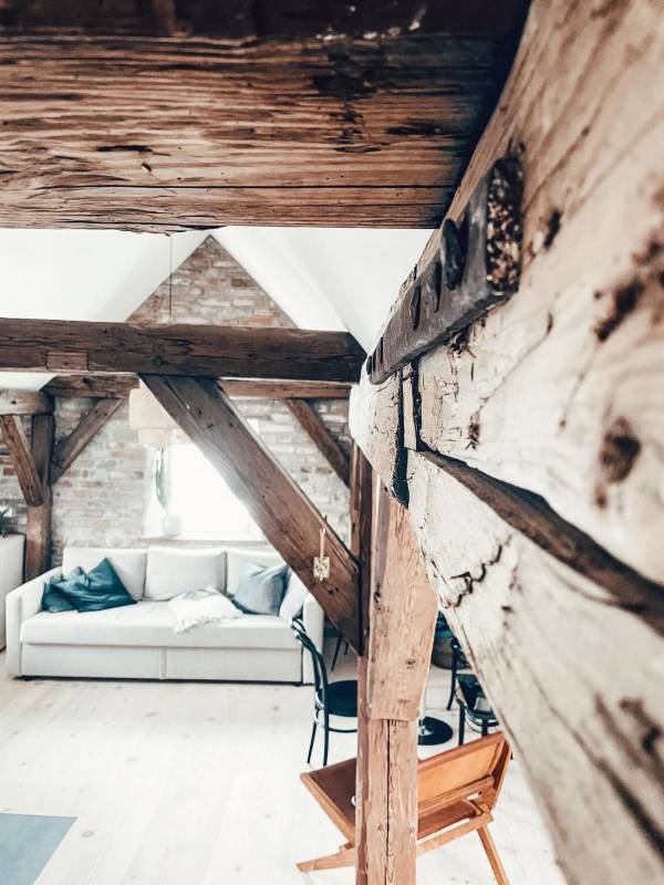 Innenansicht eines Hotelzimmers mit sichtbaren Holzträgern. Im Vordergrund ein Holzpfeiler, im Hintergrund eine Couch mit Kissen und ein Stuhl.