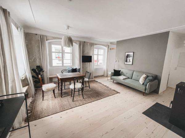 Helle Hotelzimmerdarstellung mit einem grünen Sofa, einem Holztisch und Stühlen. Ein Teppich liegt auf dem Holzfußboden. Fenster mit Vorhängen.
