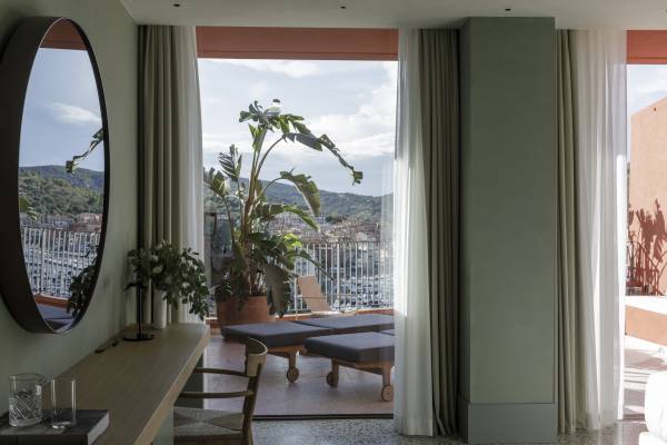 Hotelzimmer mit Schreibtisch und Stuhl, Blick durch große Fenster auf Terrasse mit Liegestühlen und Pflanzen, Meer und Hügel im Hintergrund