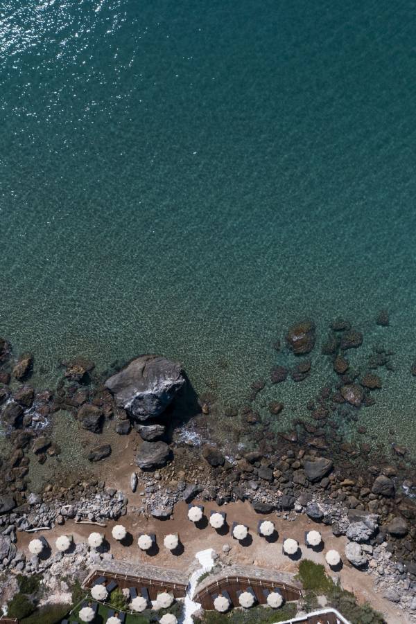 Luftaufnahme eines felsigen Küstenabschnitts mit klarem Wasser und einer Reihe von Sonnenschirmen am Strand vor einem Hotel