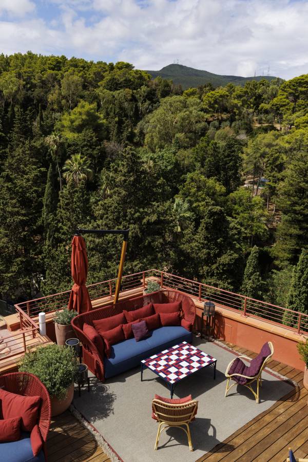 Außenbereich eines Hotels mit rotem Sofa, Stühlen und Schachbrett-Tisch auf Terrasse, umgeben von dichtem Wald und Bergen im Hintergrund