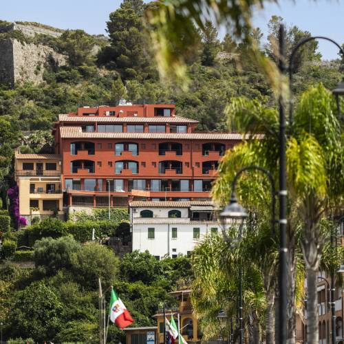 Mehrstöckiges Hotelgebäude in Rot und Beige an einem Hang mit Blick auf eine Promenade mit Palmen und Flaggen, umgeben von grüner Vegetation