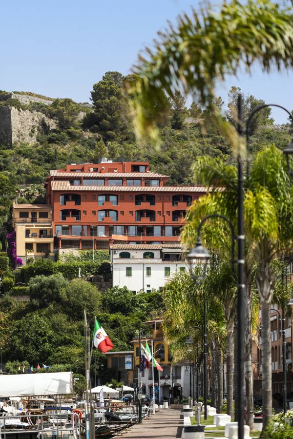 Mehrstöckiges Hotelgebäude in Rot und Beige an einem Hang mit Blick auf eine Promenade mit Palmen und Flaggen, umgeben von grüner Vegetation