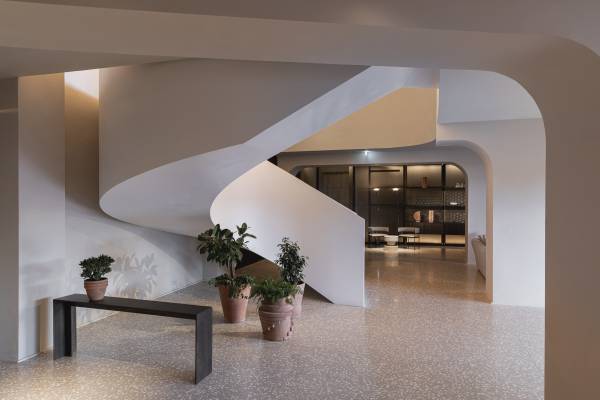 Hotelhalle mit geschwungener weißer Treppe, drei Pflanzen in Töpfen und schwarzer Bank auf hellem Boden