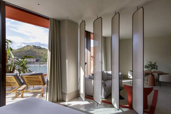 Hotelzimmer mit Blick auf Terrasse mit Liegestühlen und Berg im Hintergrund, Raumteiler aus Paneelen trennt Wohnbereich