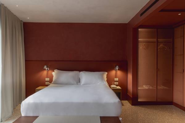 Hotelzimmer mit großem Doppelbett, weißen Bettwäsche, zwei Nachttischen mit Lampen, roter Wand und offenem Kleiderschrank