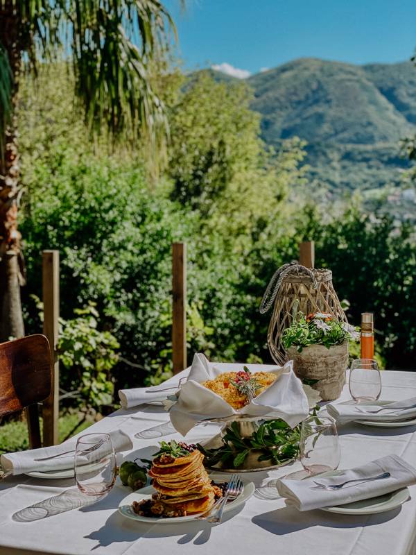 Tisch mit gedecktem Essen und weißen Servietten auf Terrasse mit Blick auf grüne Berge und blauen Himmel
