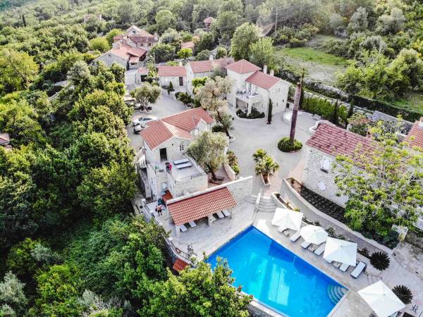 Luftaufnahme eines Hotels mit roten Ziegeldächern, umgeben von grünen Bäumen, mit einem rechteckigen blauen Swimmingpool und weißen Sonnenschirmen