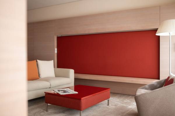 Moderne Hotelzimmeransicht mit einem hellen Sofa, einem roten Couchtisch und einem grauen Sessel. Rote Wandverkleidung hinter dem Sofa.
