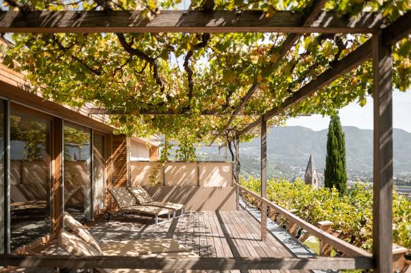 Terrasse eines Hotels mit Holzrahmen und Weinreben, Blick auf Berge und einen Kirchturm im Hintergrund.