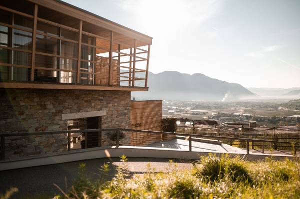 Moderne Hotelanlage mit Holz- und Steinfassade, umgeben von einer grünen Landschaft und Bergen im Hintergrund. Klare Sicht auf das Tal.