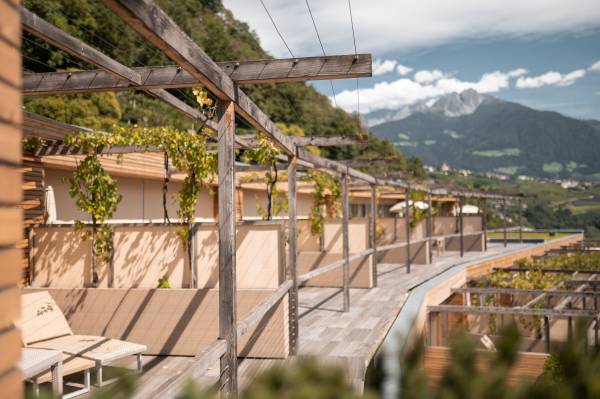 Blick auf eine Hotelanlage mit Holzpergolen und Weinreben. Im Hintergrund sind Berge und eine grüne Landschaft sichtbar.